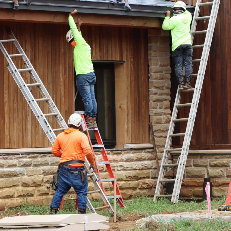 Two workers on ladders reach up to a building's eave while a third stands below