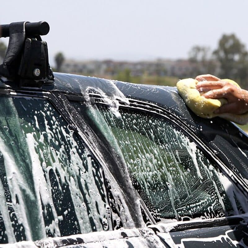A hand washing a dark vehicle with a yellow mitt, soap running down the side windows
