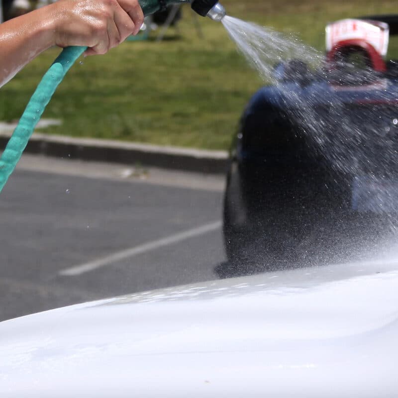 A garden hose rinsing soap off a white hood, a dark car behind it