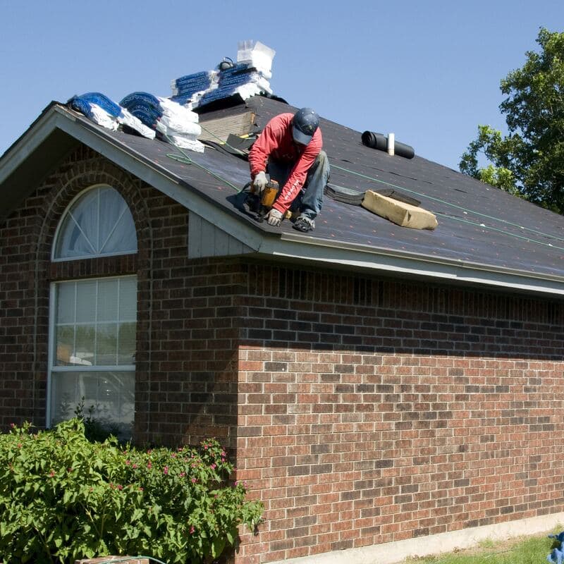 A roofer with a nail gun on a brick home's roof, underlayment down and bundles stacked at the ridge