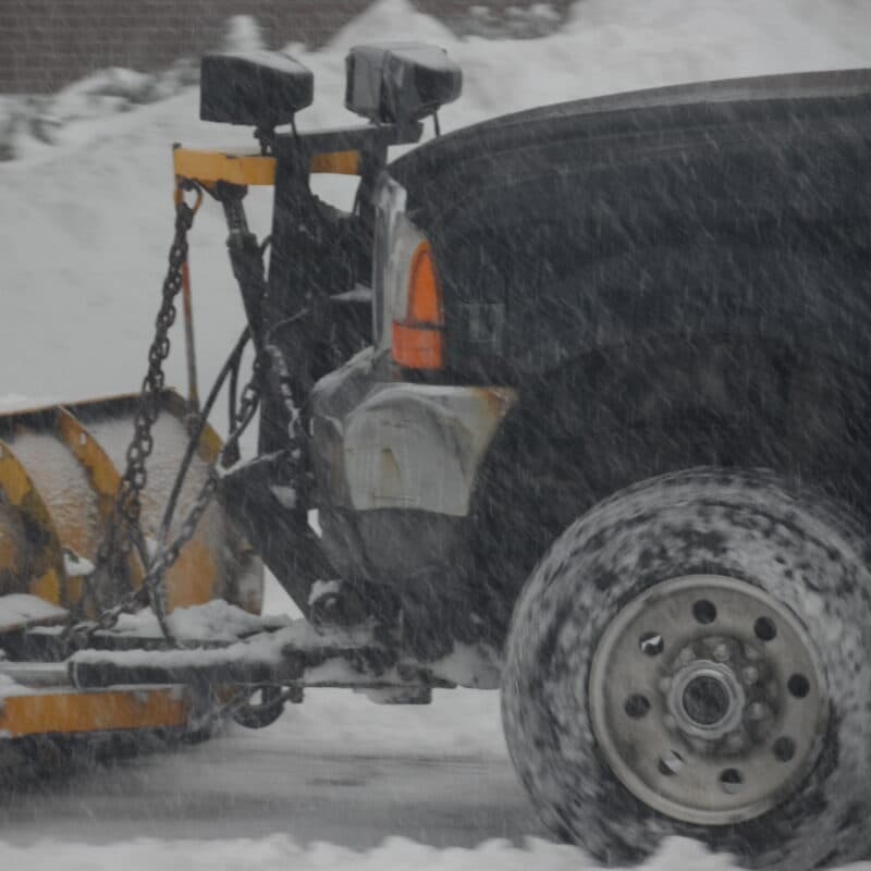 A pickup truck with a plow blade pushing snow across a lot during a storm