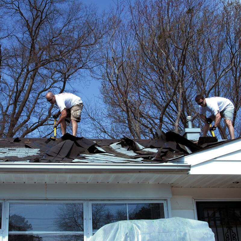 Two roofers on a low roof prying up old shingles, torn shingles piled along the eave