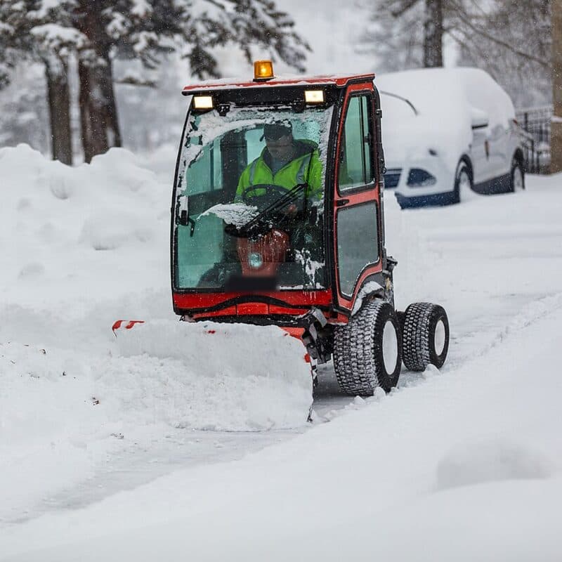 An operator in a hi-vis vest drives a cab-enclosed walk machine with a blade along a snowy sidewalk